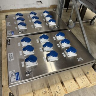 Kaksi ruostumattomasta teräksestä Eiskon valmistamaa pistorasiakeskusta lavan päällä varastotilassa. Kummassakin keskuksessa on yhdeksän sinistä 16 A -teollisuuspistorasiaa.