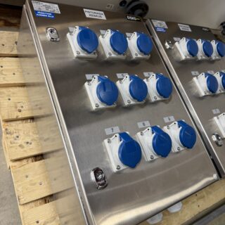 Kaksi ruostumattomasta teräksestä Eiskon valmistamaa pistorasiakeskusta lavan päällä varastotilassa. Kummassakin keskuksessa on yhdeksän sinistä 16 A -teollisuuspistorasiaa.