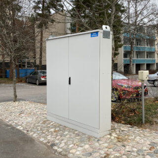 Harmaa Eiskon valmistama sähkön pääkeskus ulkotilassa kivetyksellä, taustalla kerrostaloja ja pysäköityjä autoja.